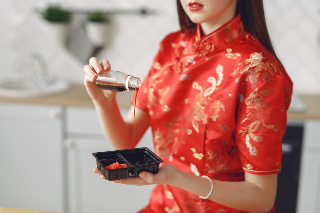 Unrecognizable lady in vivid red dress holding disposable capacity for sauce with ginger and pouring soy sauce from bottle while eating in modern kitchen