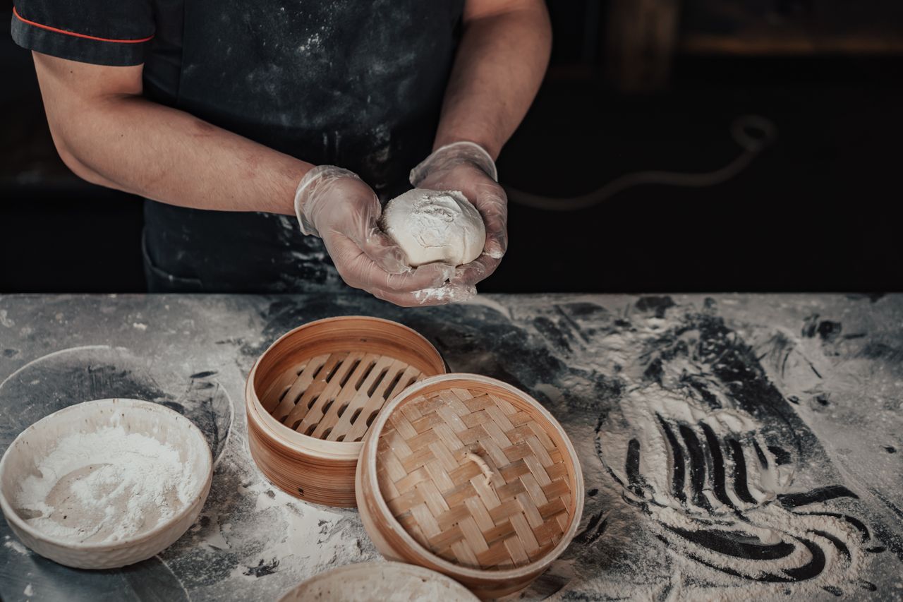 A chef carefully handcrafts baozi dough with a bamboo steamer on a flour-dusted table.